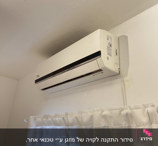 מזגן קיר לבן מותקן מעל חלון עם וילון לבן